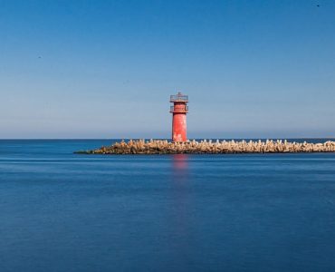 A vibrant red lighthouse stands on a tranquil coastline in Ras El-Bar, Egypt, under a clear blue sky.