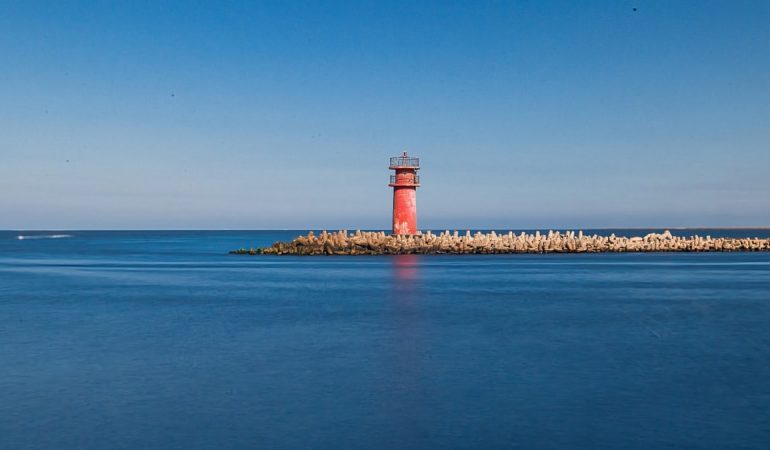 A vibrant red lighthouse stands on a tranquil coastline in Ras El-Bar, Egypt, under a clear blue sky.